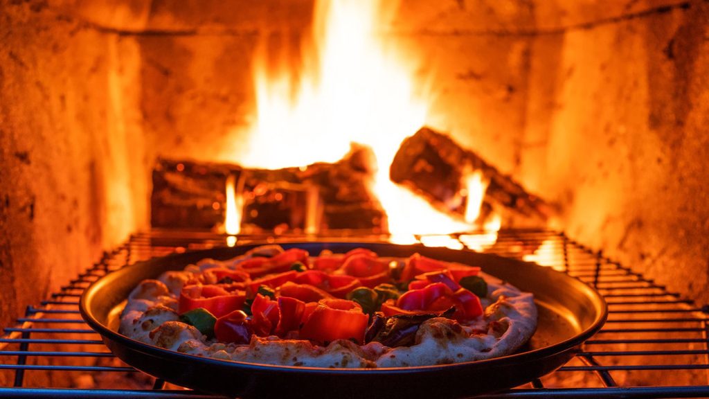Pizza in cottura su una griglia davanti alle fiamme di un forno a legna